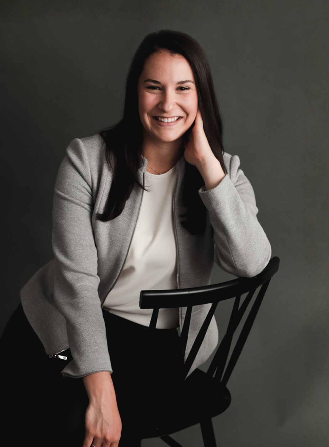 Woman in a light gray blazer smiles, sitting on a black chair against a gray backdrop.