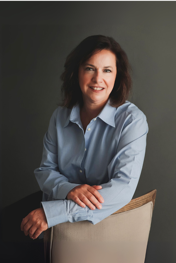 Woman with brown hair, wearing a blue button-down shirt, smiles, leaning on the back of a chair, against a gray wall.