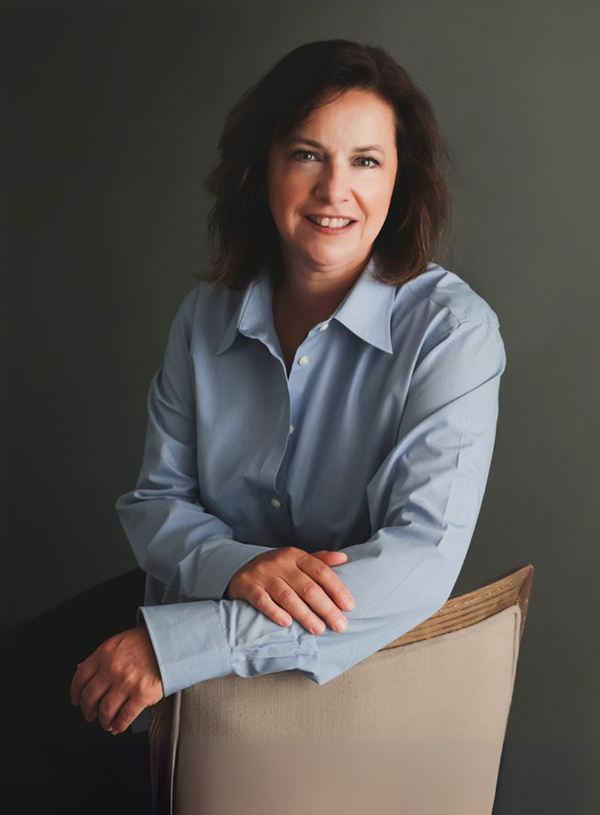 Woman in blue button-down shirt smiles, leaning on the back of a light chair against a gray background.