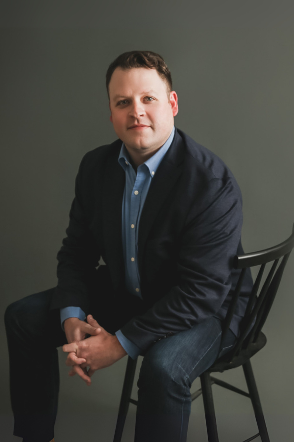 Man in blazer and jeans sits on a chair, looking forward against a gray backdrop.