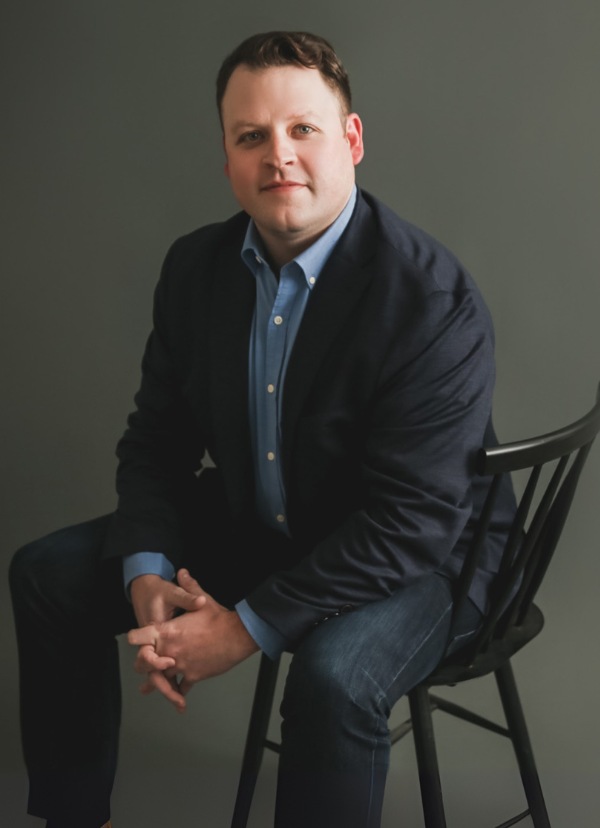 Corey Ernisse seated on a black chair, wearing a navy blazer over a blue shirt