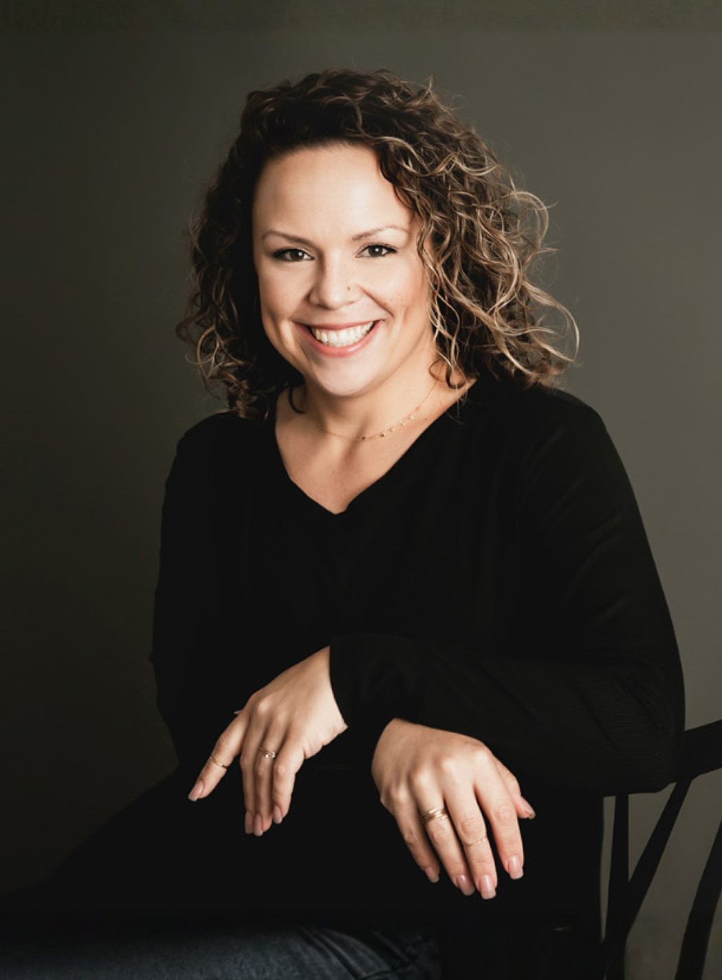Woman with curly hair smiles, seated, wearing a black sweater, against a gray background.