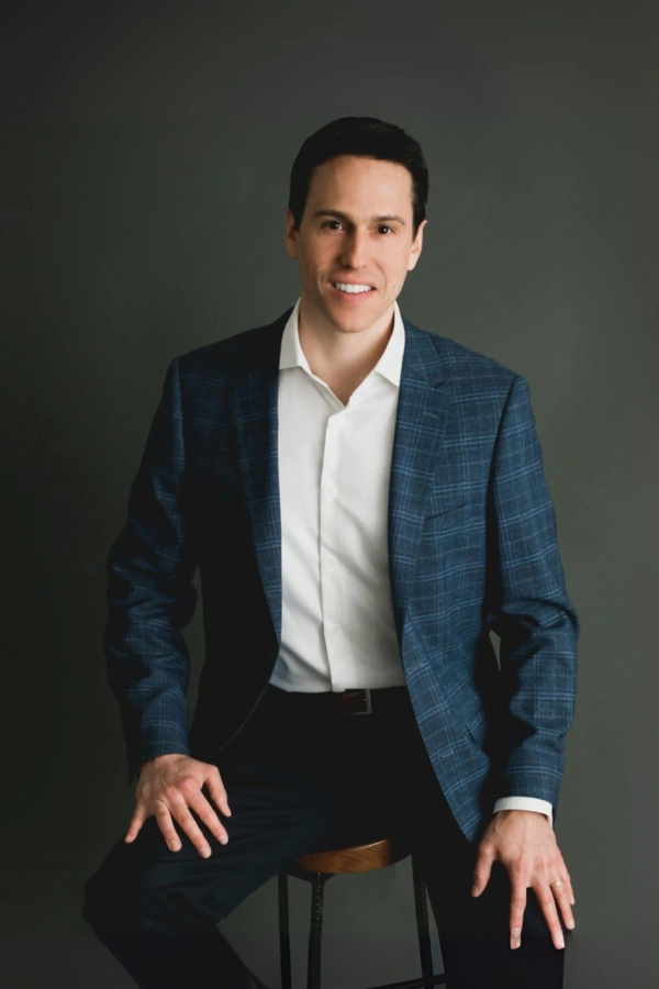 Man in blue blazer and white shirt smiles, seated on stool against a gray background.