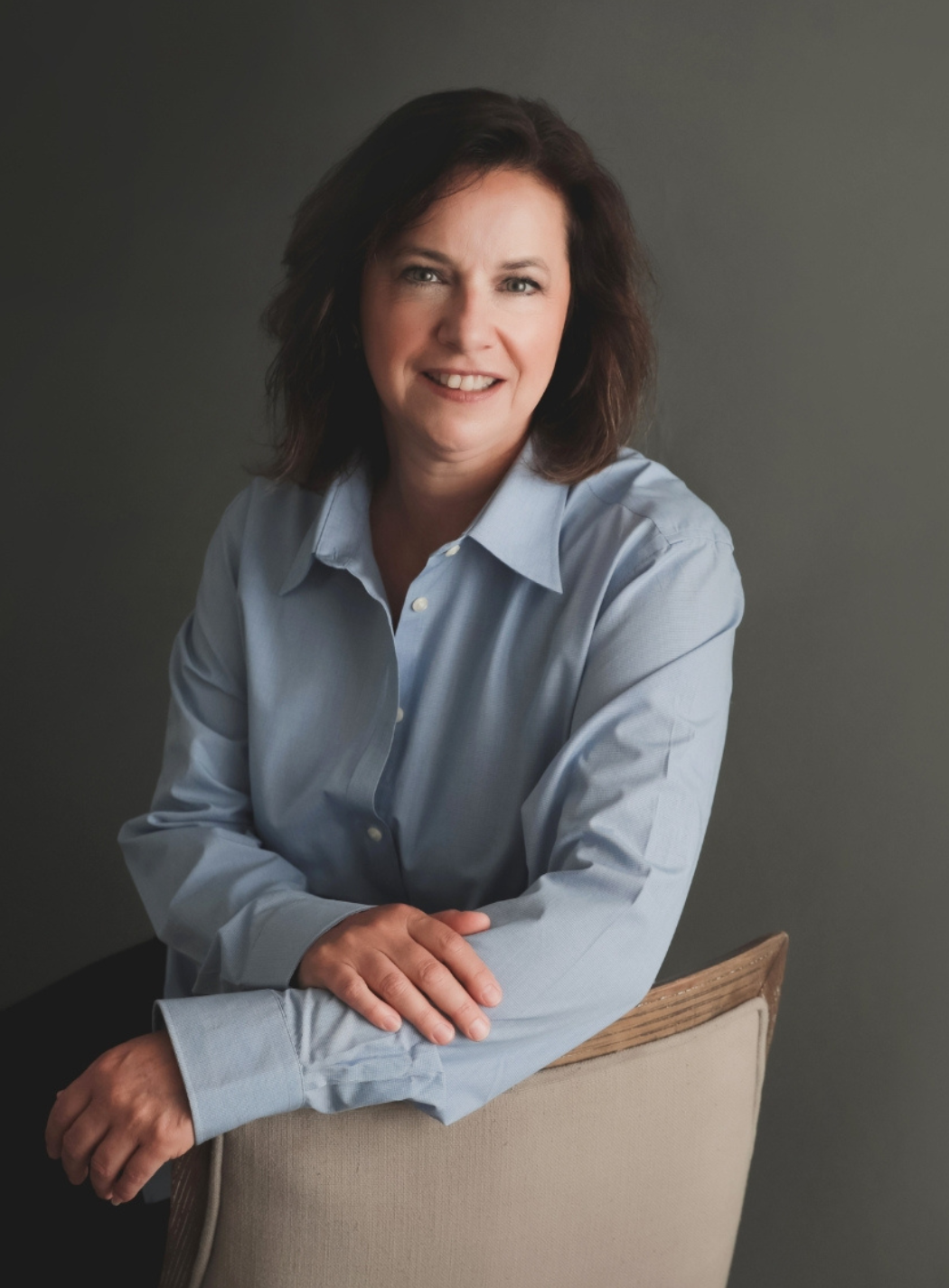 Woman in light blue button-down shirt smiles, leaning on chair. Gray background.