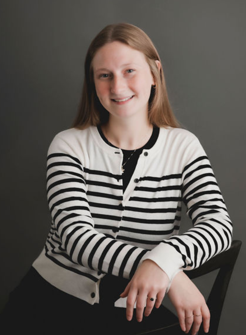 Woman smiling, wearing a striped cardigan, seated in front of a gray backdrop.
