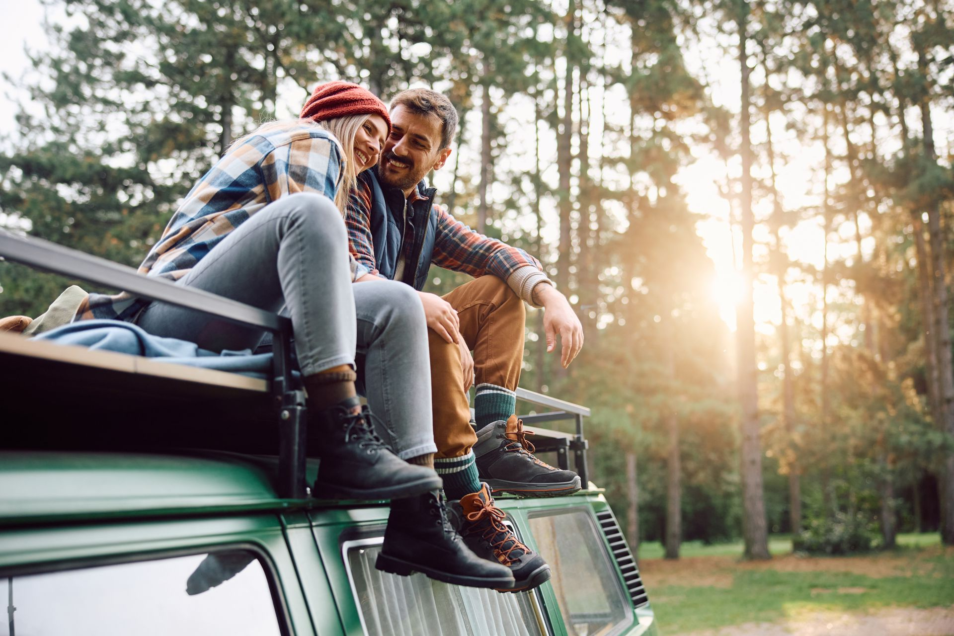 Couple sits on top of green van in a forest, sun shining through trees.