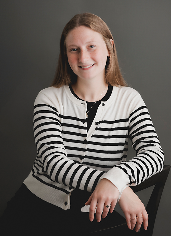 Woman smiling, wearing a striped cardigan, seated, hands on chair back, against a grey backdrop.
