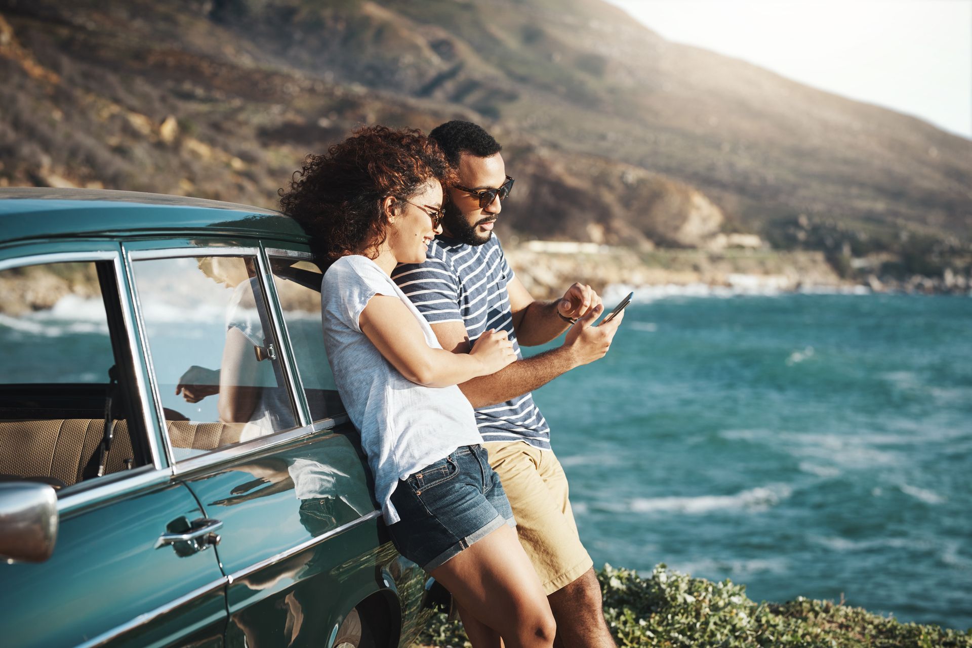 Couple by car, looking at tablet near ocean; mountains in background.