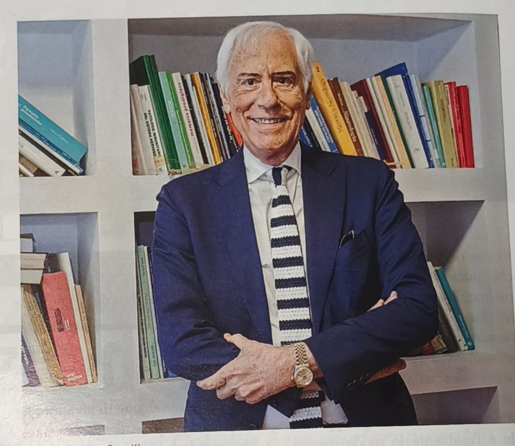 Un uomo in abito blu scuro è in piedi davanti a una libreria, sorridente con le braccia incrociate.