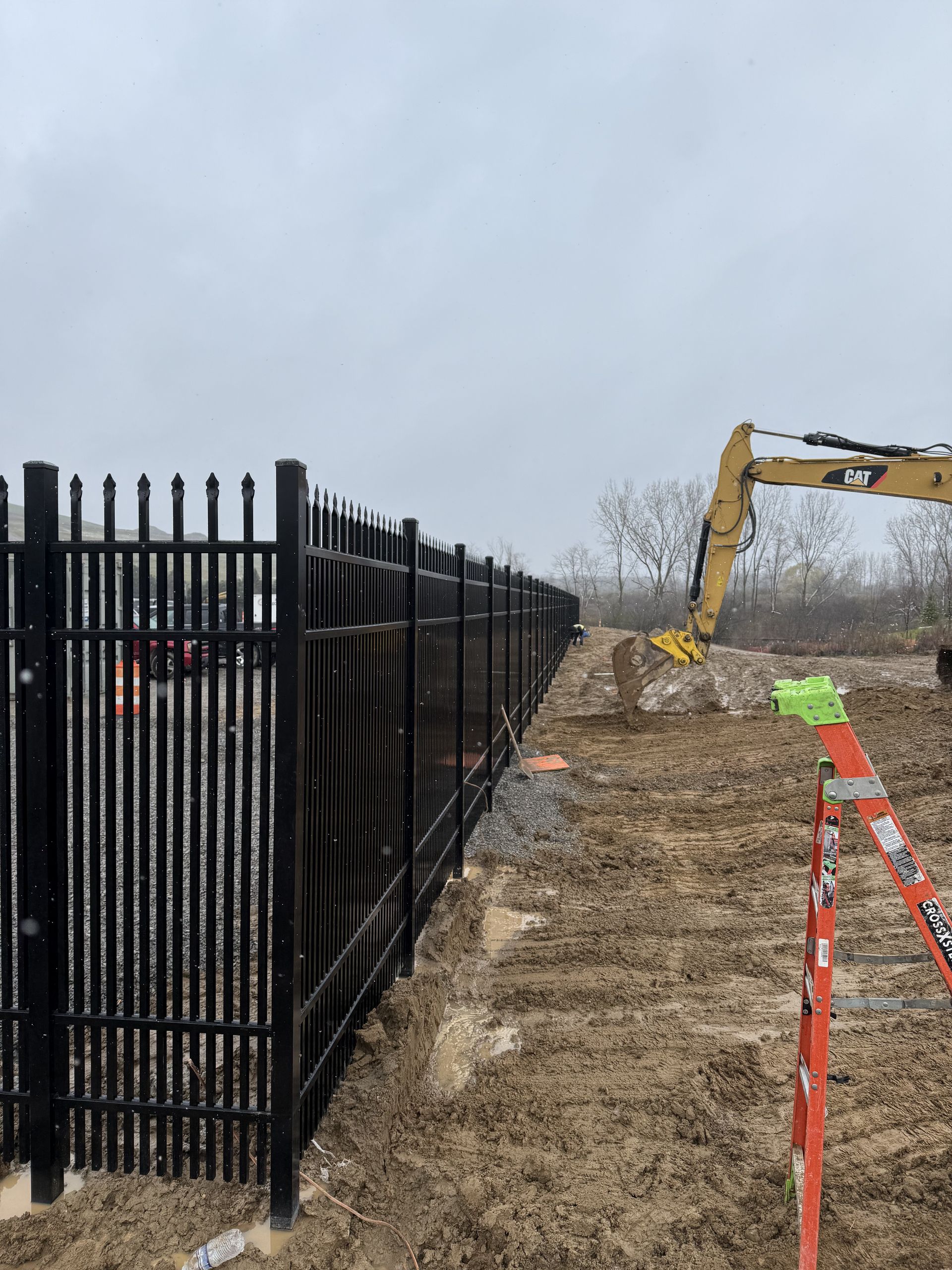 Black metal fence along muddy ground with construction equipment in the background under an overcast sky.