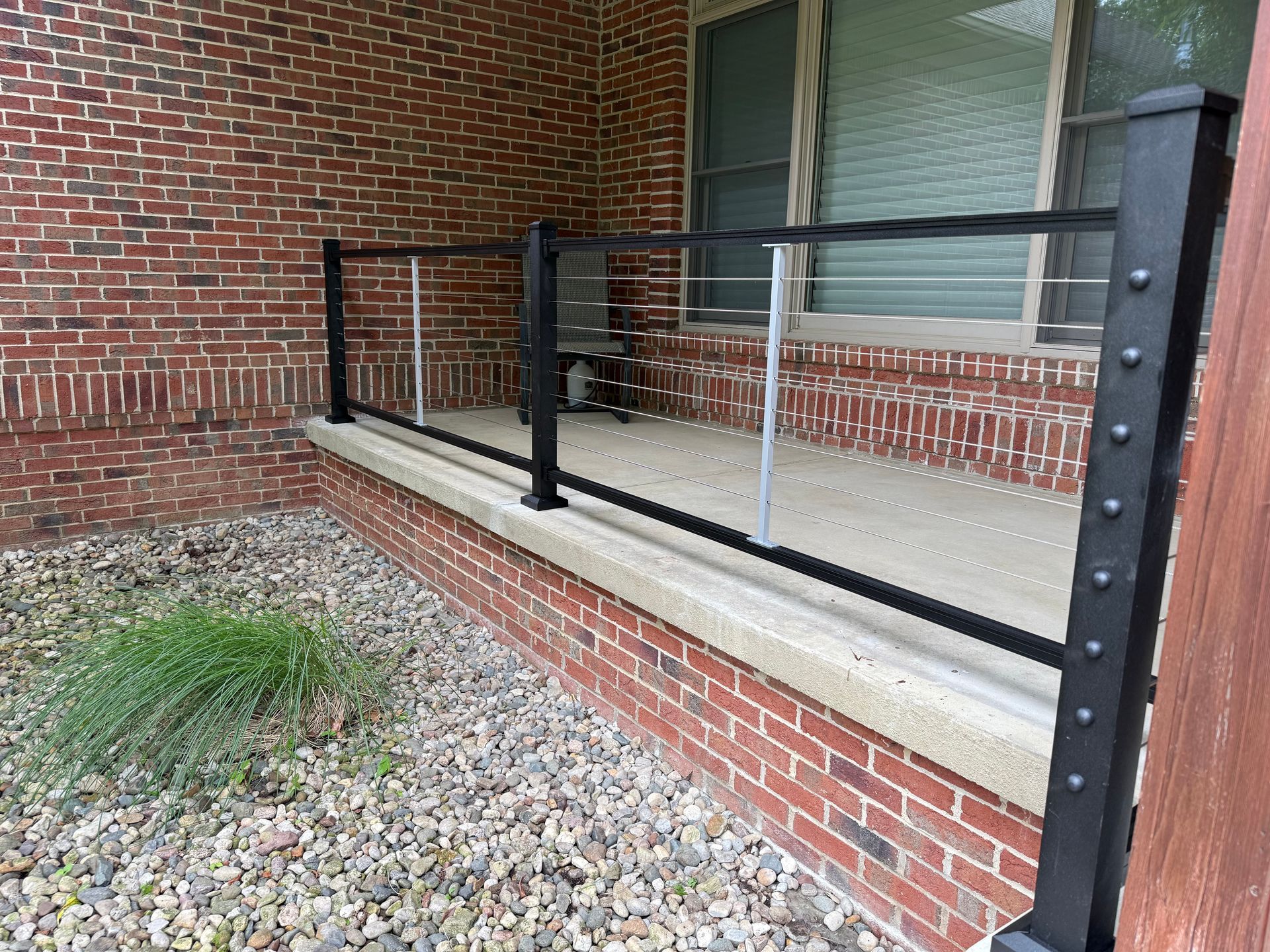 Black metal railing with stainless steel cables on a brick porch, in front of a window.