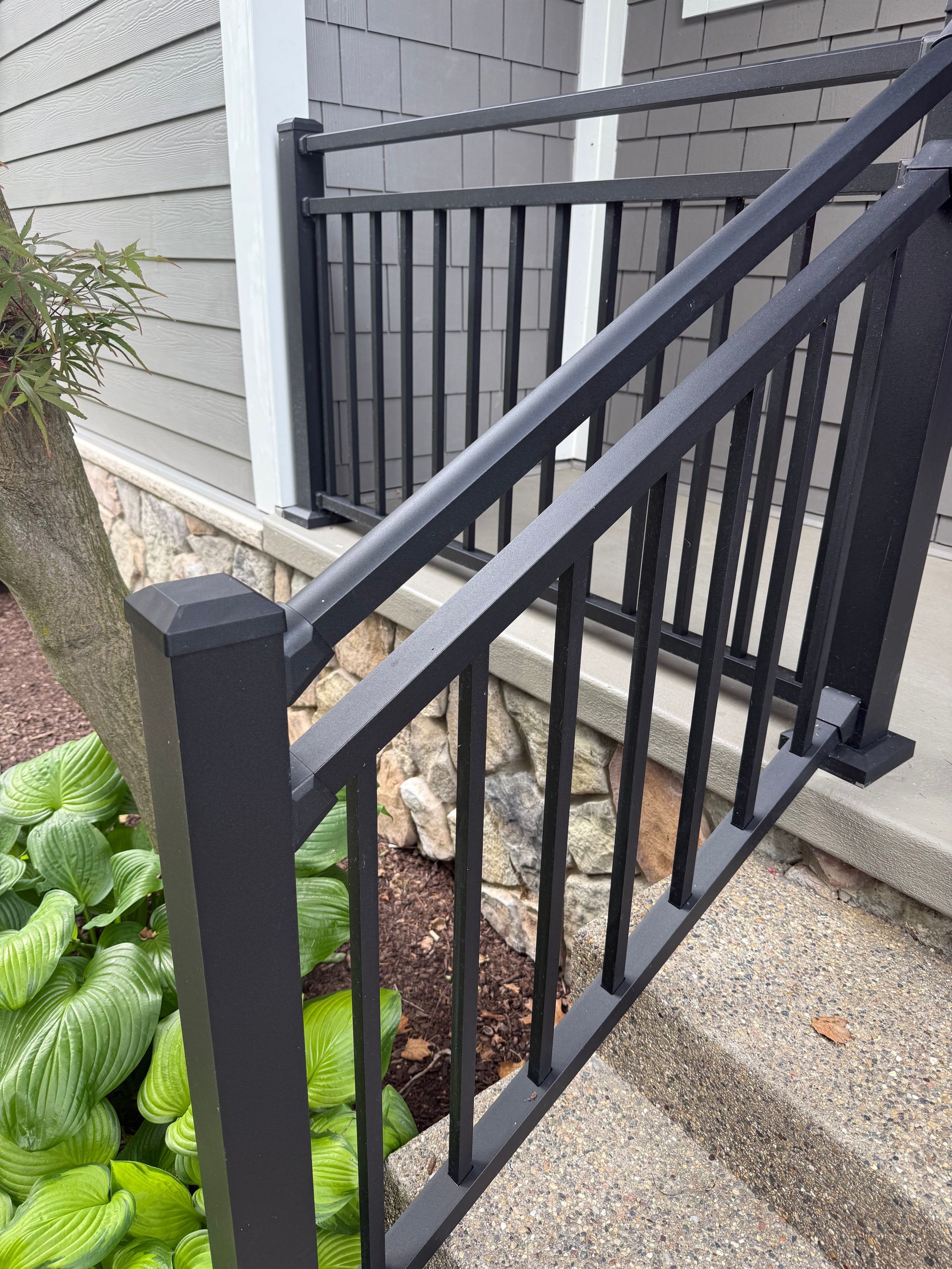 Black metal railing on concrete steps, next to a stone wall and plants. Gray siding background.
