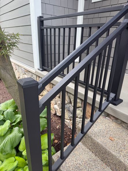 Black metal railing on concrete steps, next to a stone wall and plants. Gray siding background.