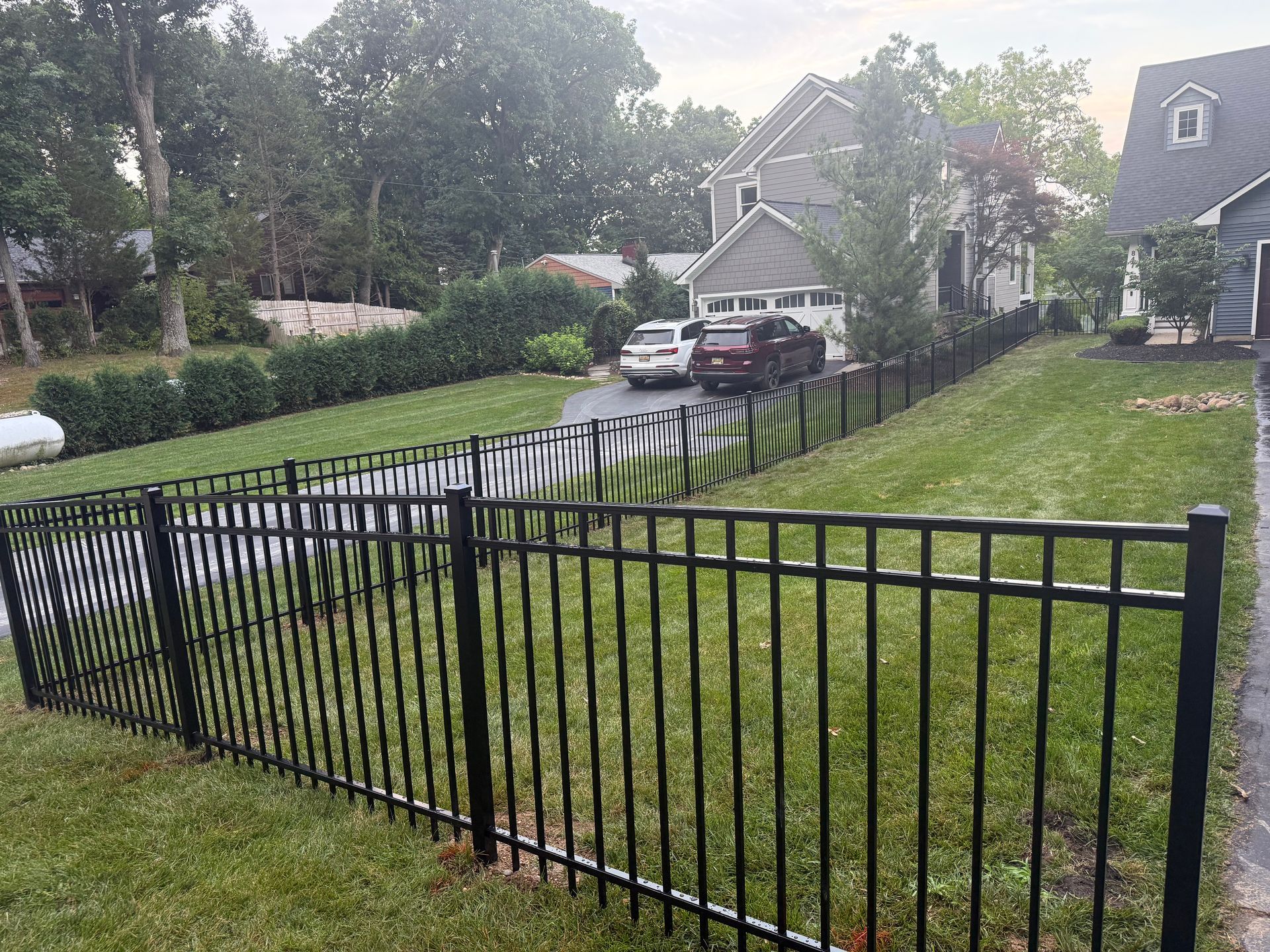 Black metal fence bordering a green lawn, leading to driveways and houses with trees in the background.
