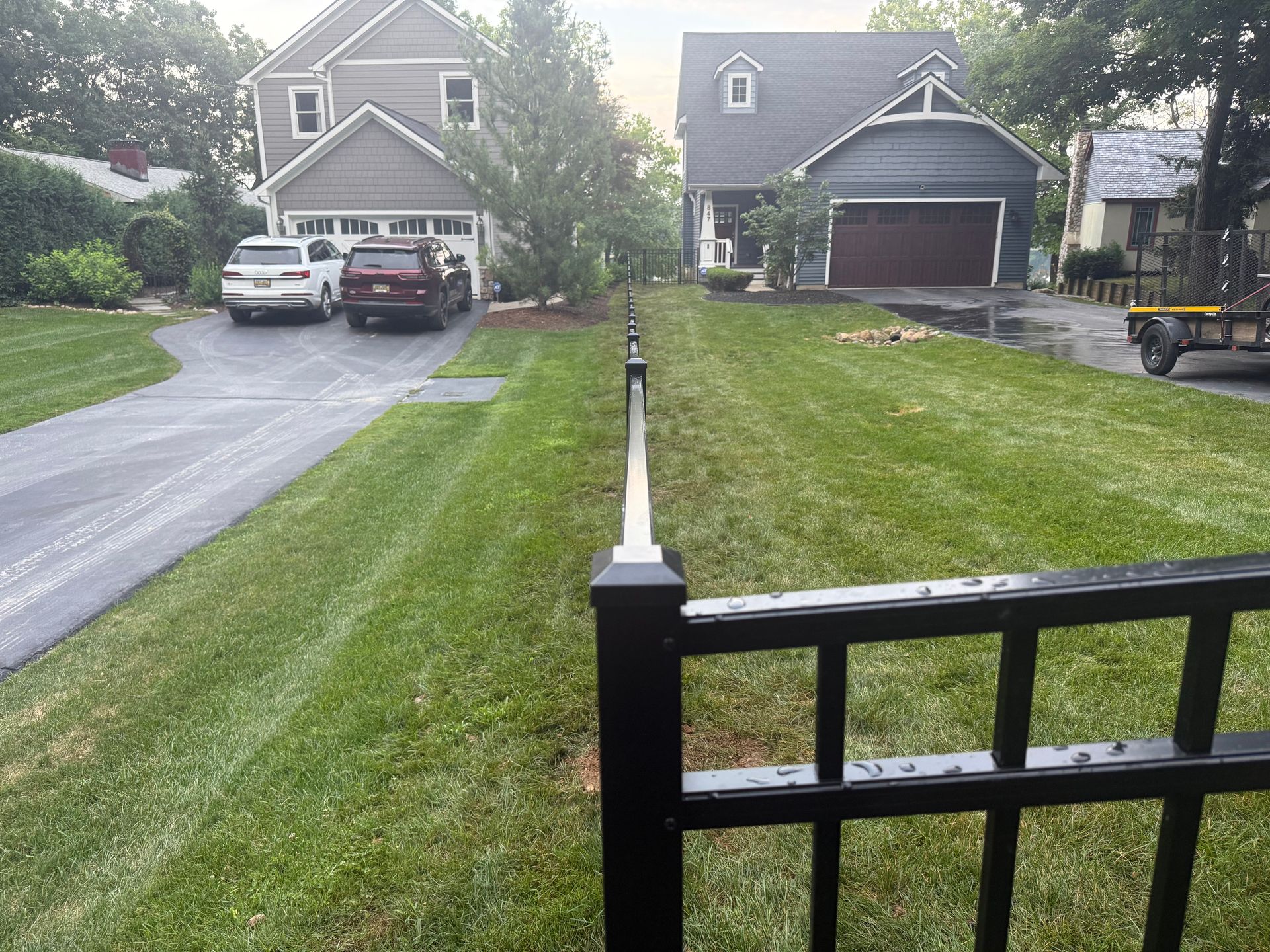 A black fence lines the grassy yard between two houses with cars parked in the driveways.