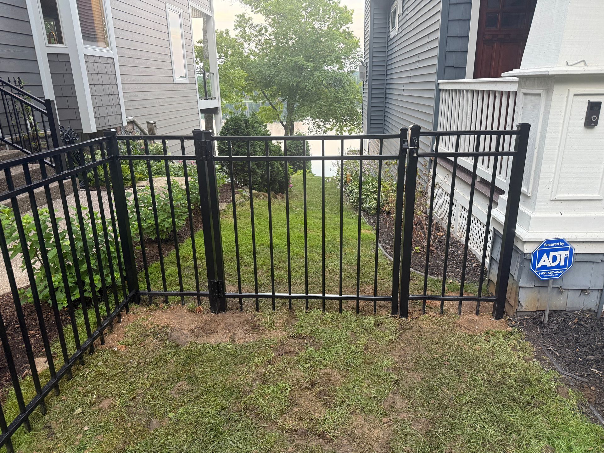 Black metal fence with gate, enclosing a small grassy yard between two houses.