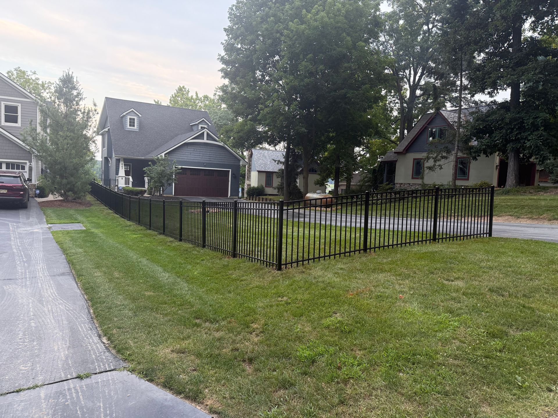 Lawn and house with a black picket fence in front. Green grass and trees under a cloudy sky.