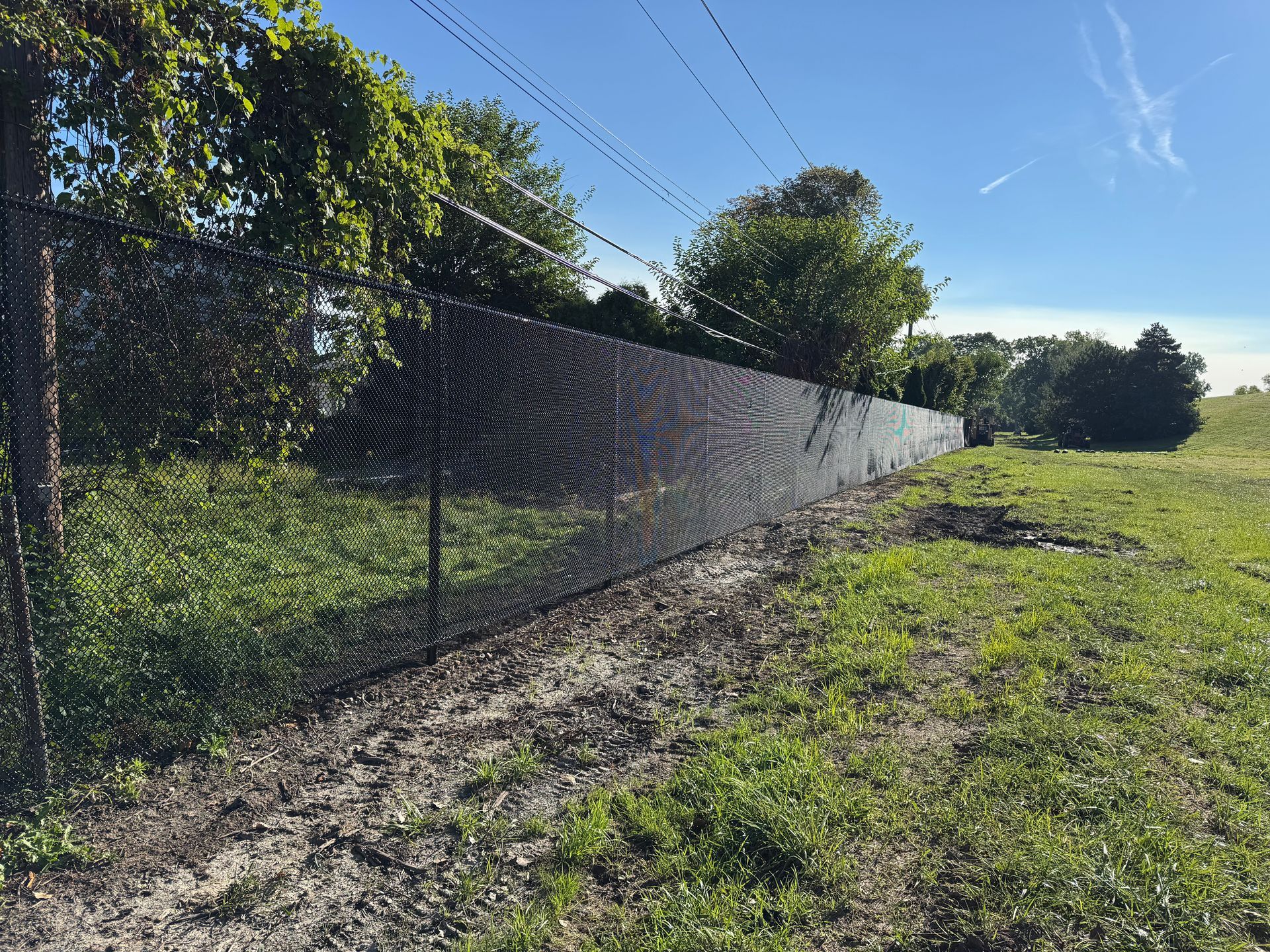 Black privacy screen covering chain-link fence in field, trees and blue sky.
