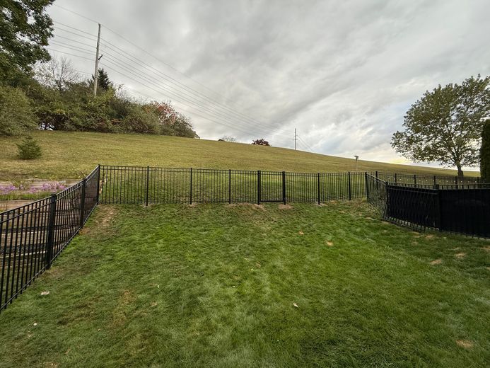 Green grassy backyard with a sloped hill and black fence under a cloudy sky.