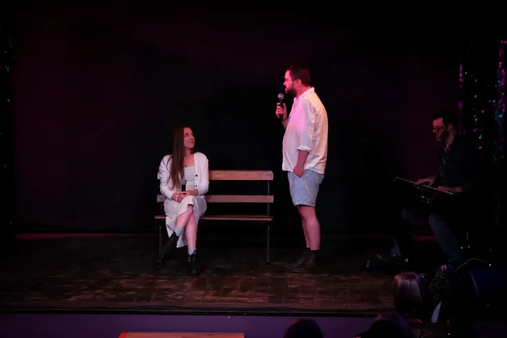 Two people on a dimly lit stage, one seated on a bench and one standing nearby.