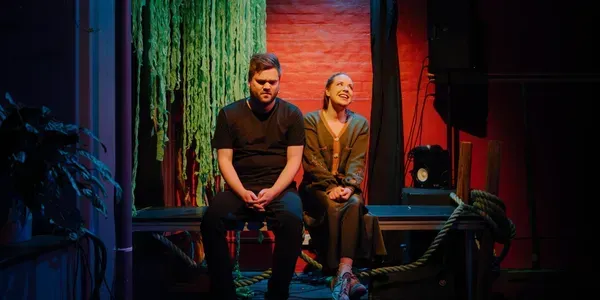 Two people seated on a bench in colorful stage lighting beside hanging plants and a brick wall.