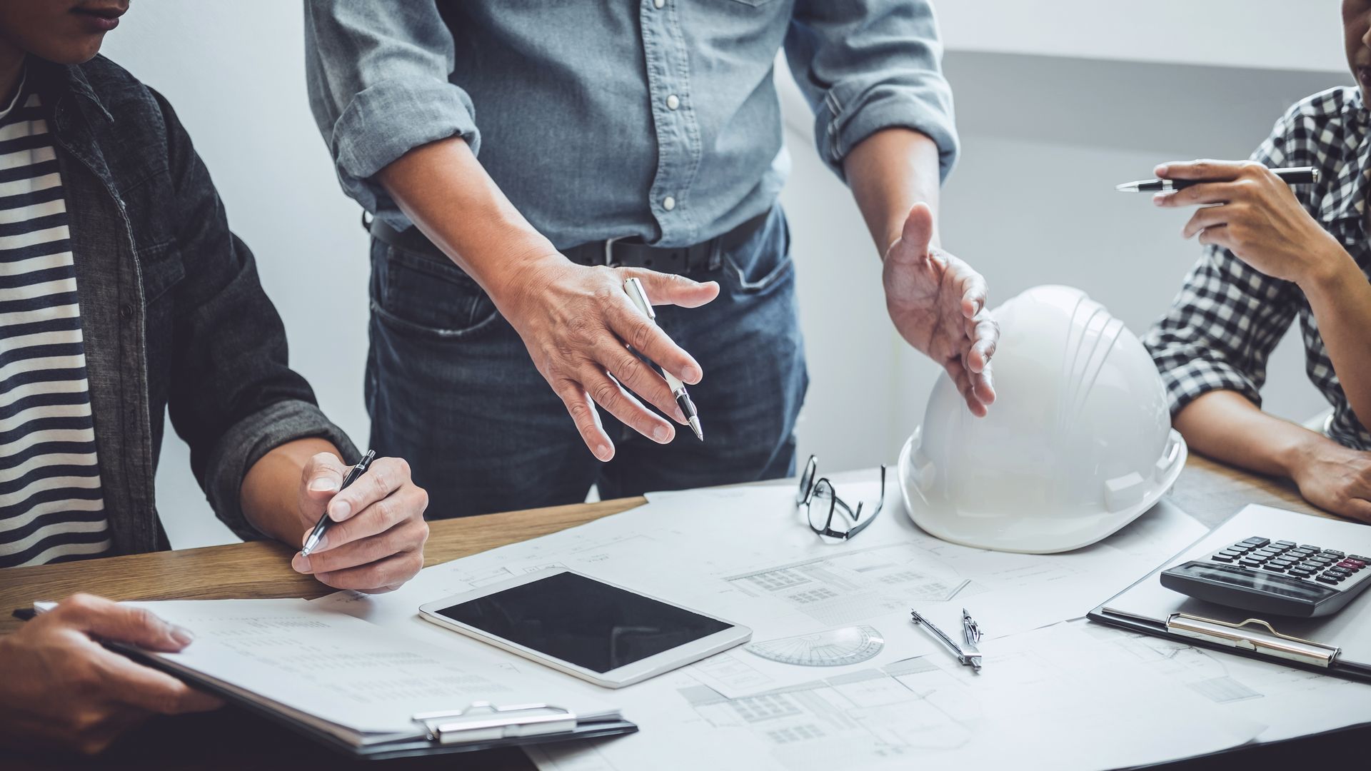 Group of people reviewing blueprints at a table, discussing a project plan. A white hard hat sits nearby.
