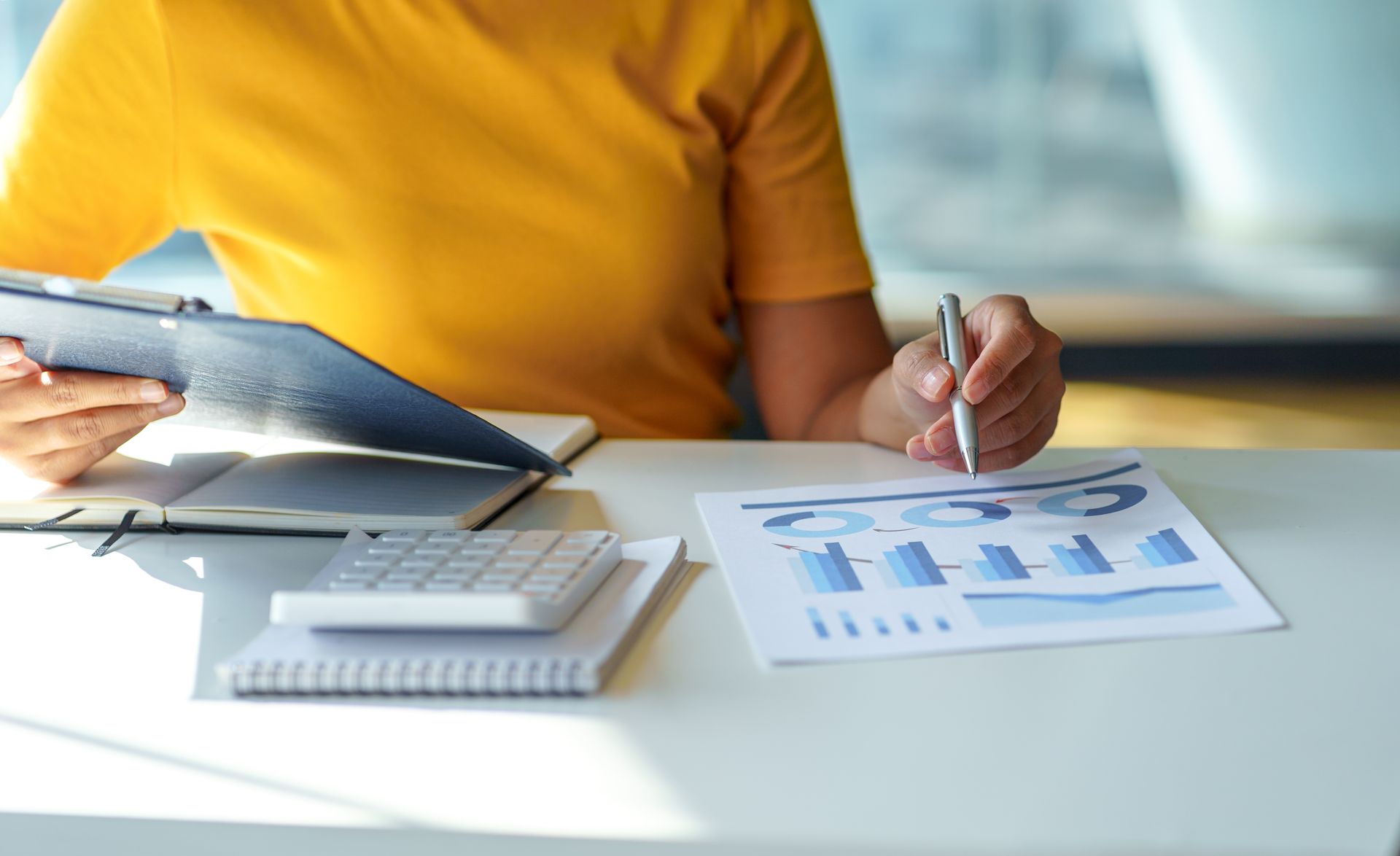 Person in yellow shirt reviewing financial charts at a desk with a calculator and notepad.
