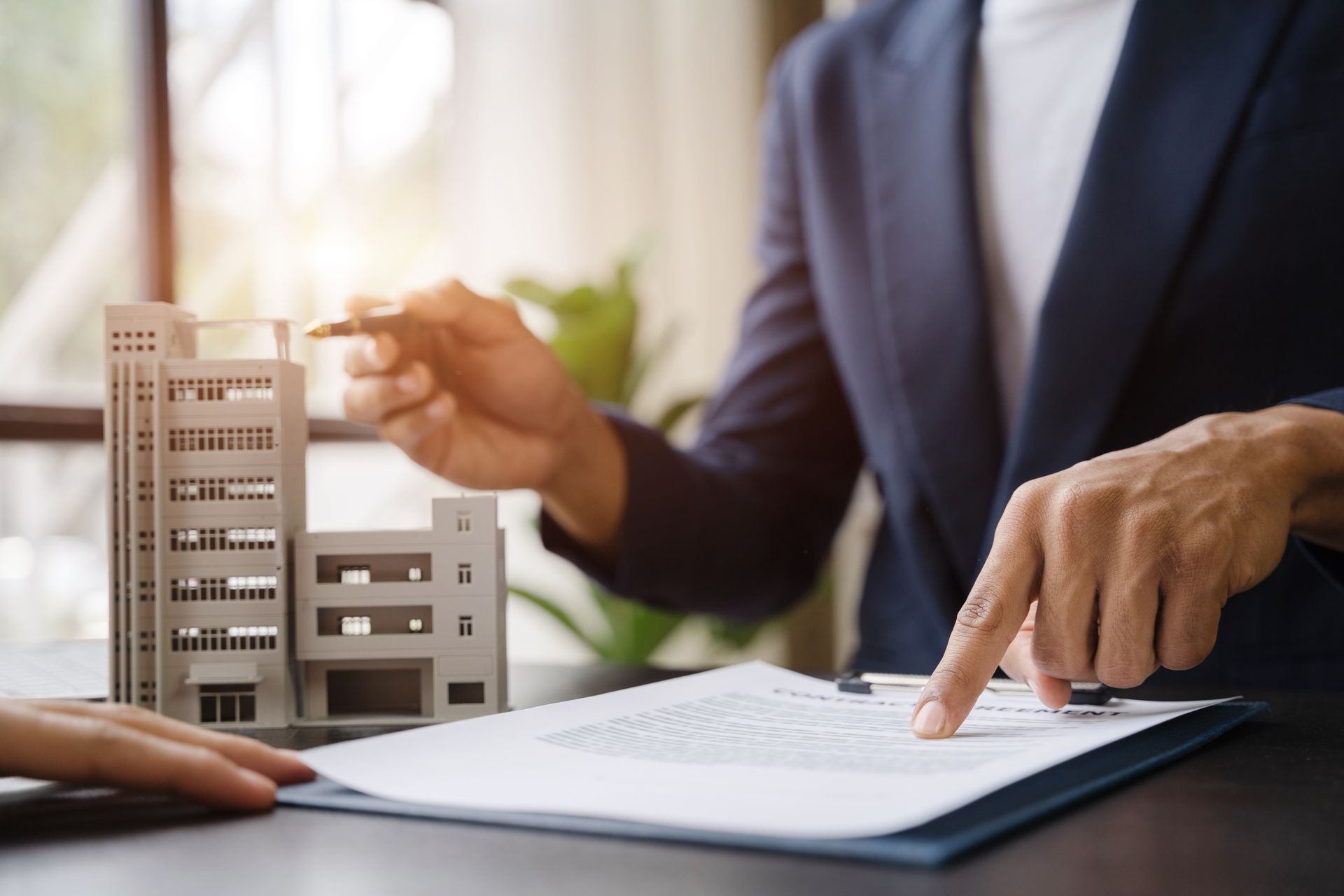 Person pointing to a document, small building model on desk, possibly real estate deal.