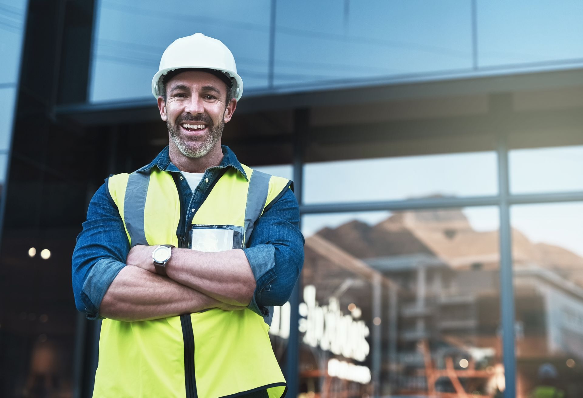 Construction worker wearing a hard hat and safety vest smiling with arms crossed in front of a modern building.