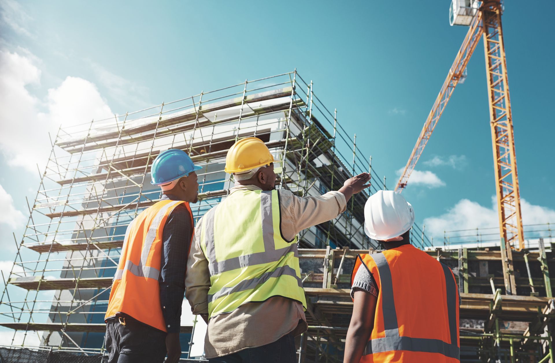 Construction workers in safety vests and hard hats look at a building under construction, crane in background.