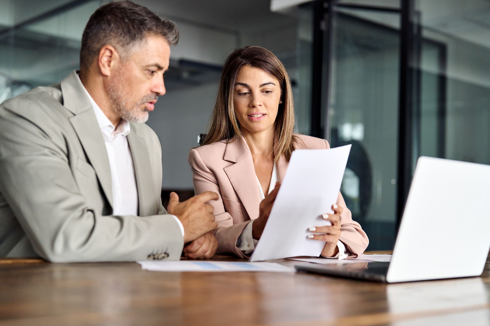 Man and woman reviewing papers together at a table, both looking at documents, laptop in view.