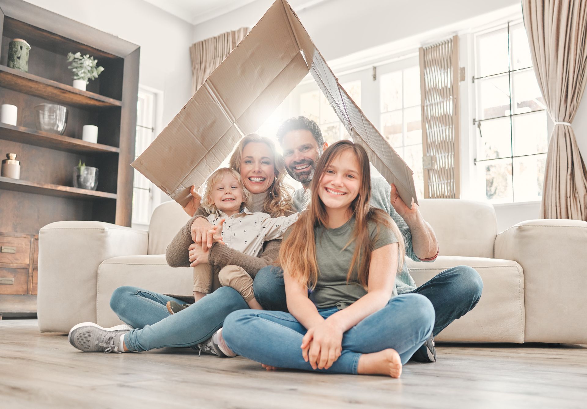 Family under a cardboard house; smiling, sitting on floor in front of a couch, indoor setting with window.