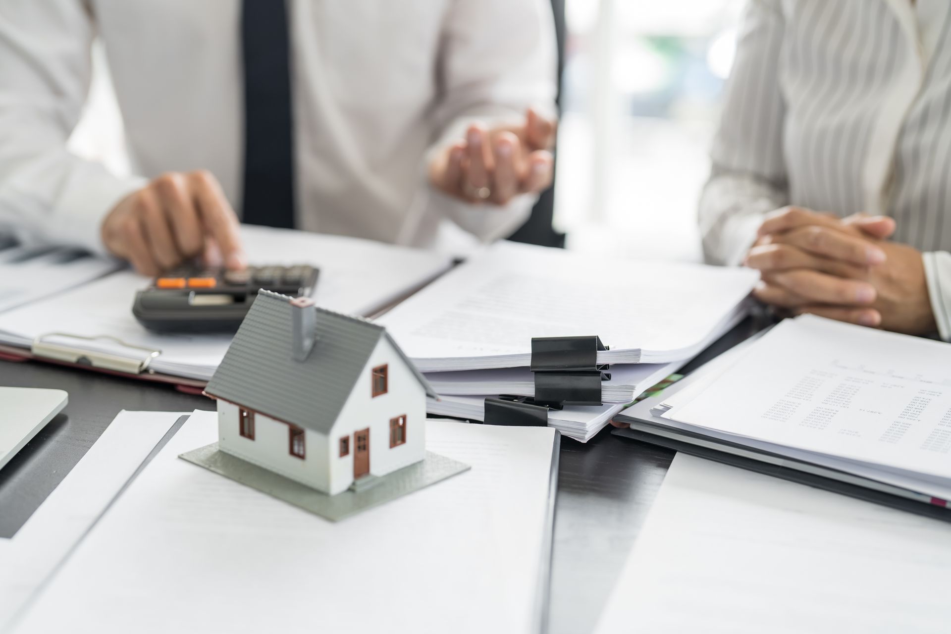 Miniature house on documents; two people in business setting, one using a calculator.