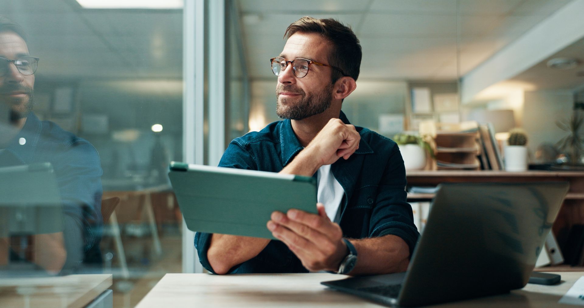 Man with glasses looks out window while holding a tablet, sitting at a desk with a laptop.