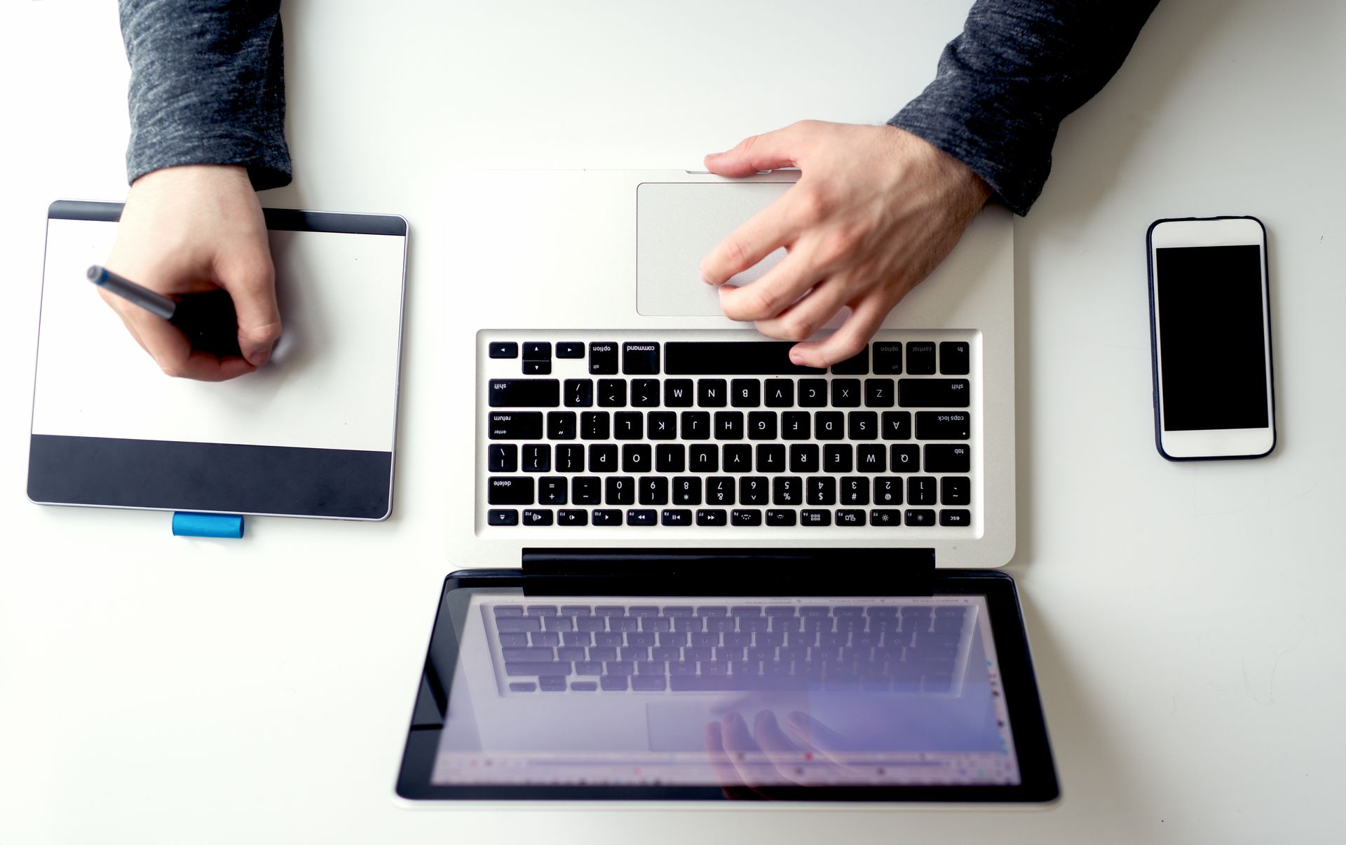 Hands using laptop, tablet, drawing tablet, and smartphone on a white surface.