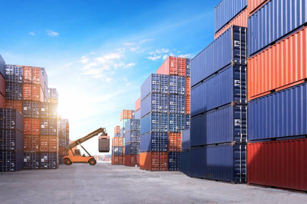 Shipping containers stacked in a port; a forklift loads a container under a bright sky.