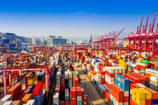 Cargo containers stacked in a bustling port; red cranes, city skyline in background, blue sky.
