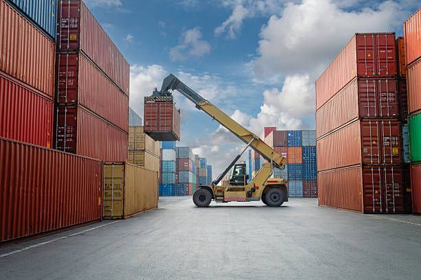 Forklift lifting a shipping container in a busy port with many colorful containers stacked high under a cloudy sky.
