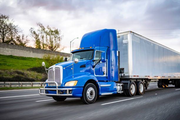 Blue semi-truck driving on a highway, hauling a silver trailer. Cloudy sky in background.