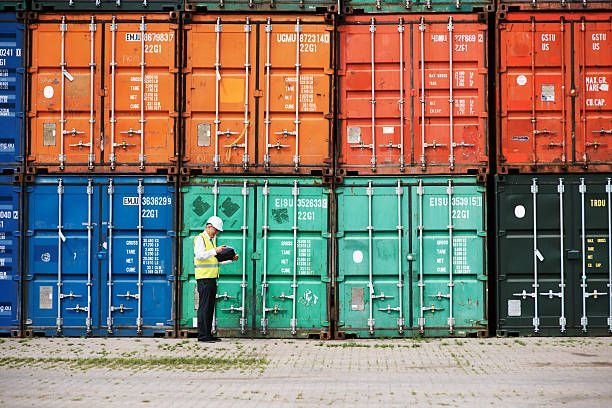 Man in hard hat and vest reviews a tablet in front of stacked, colorful shipping containers.