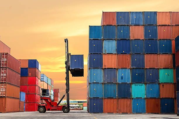 Forklift lifting a blue shipping container in a dockyard, stacked containers in the background, orange sky.