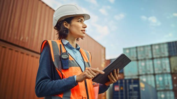 Woman in hard hat and safety vest, holding tablet, surveys shipping containers.