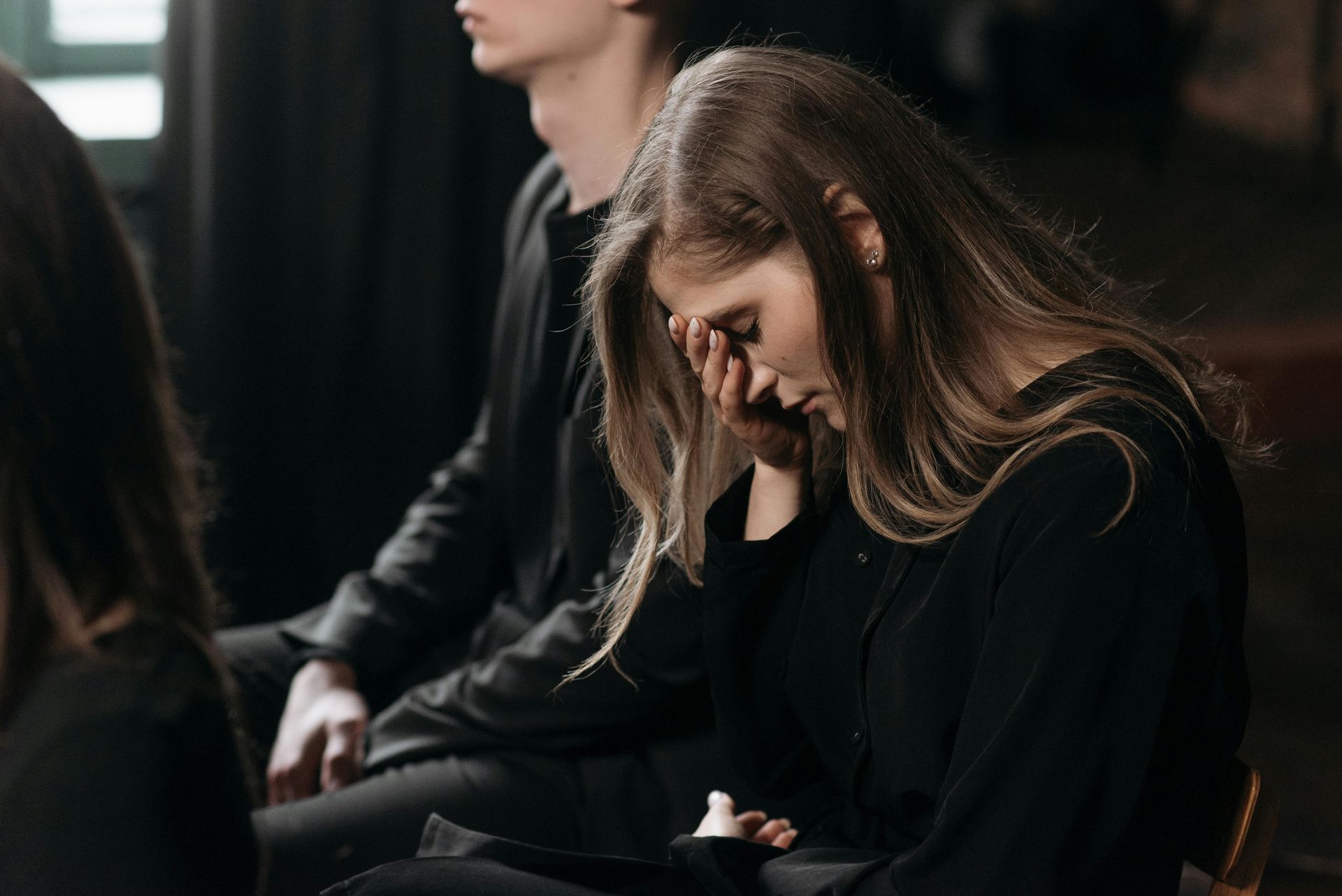 Seated people in dark clothing, one woman looking down with her hand on her face in a dim room