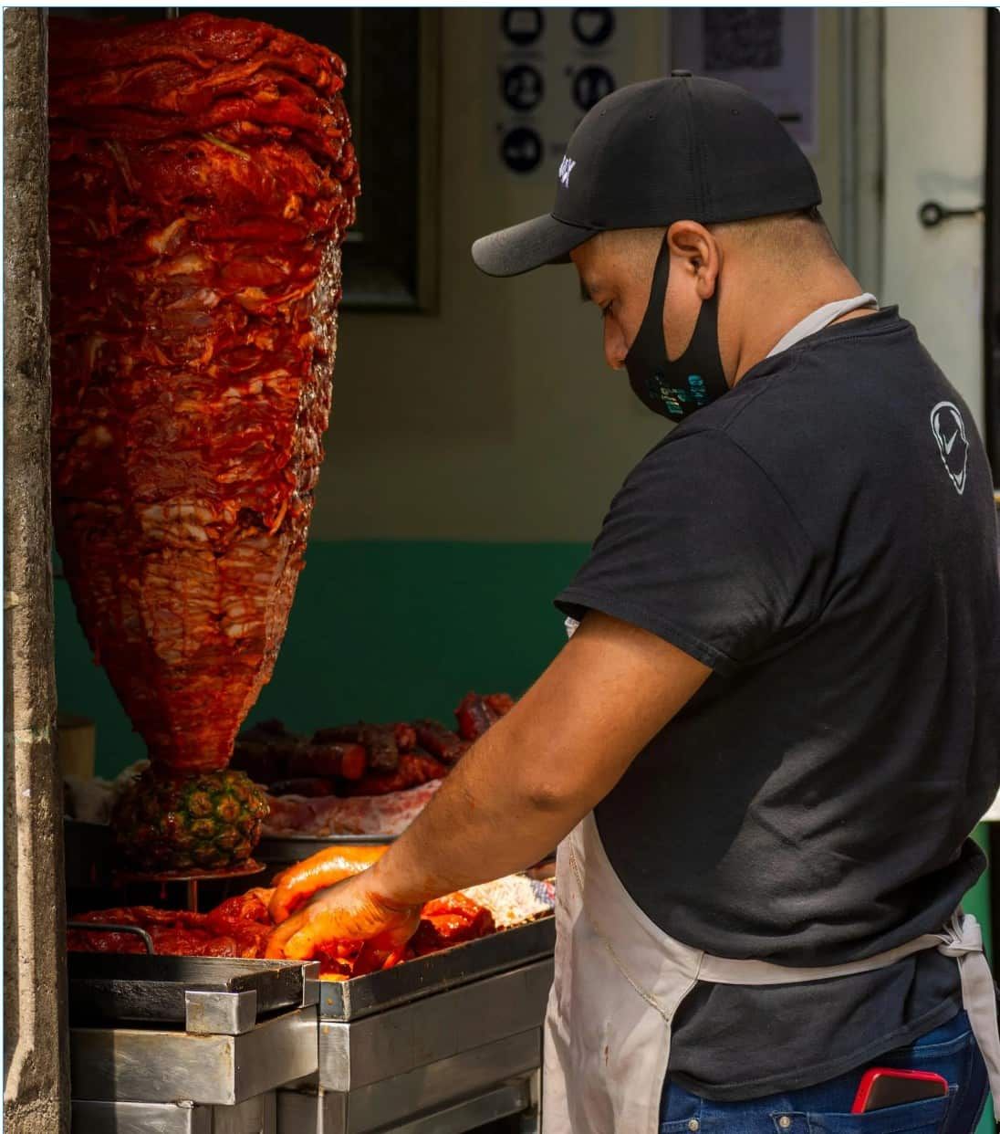 Man in black cap and mask prepping meat from a vertical rotisserie.