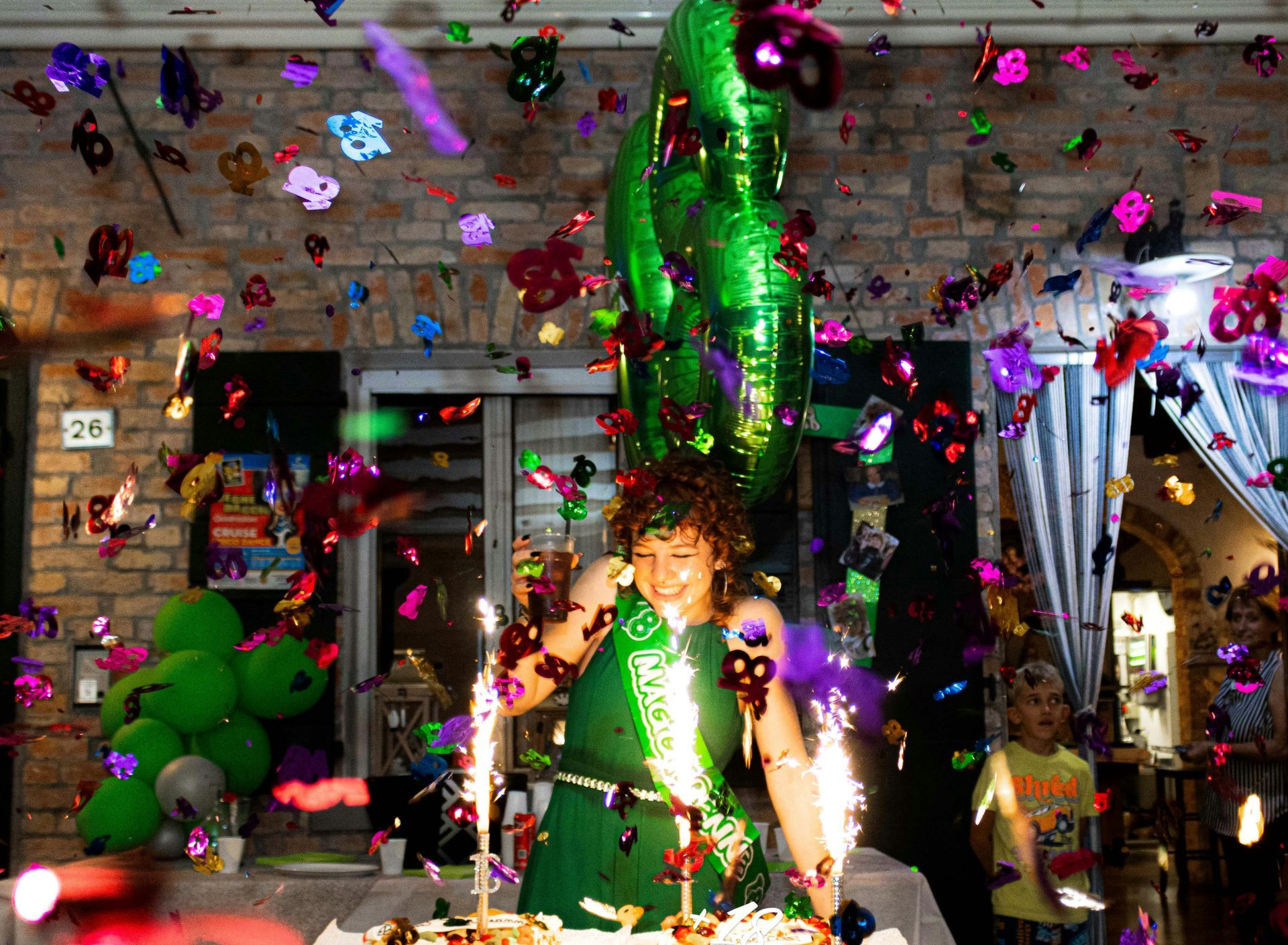 Person in green outfit celebrates with confetti and sparklers near a cake and balloons.