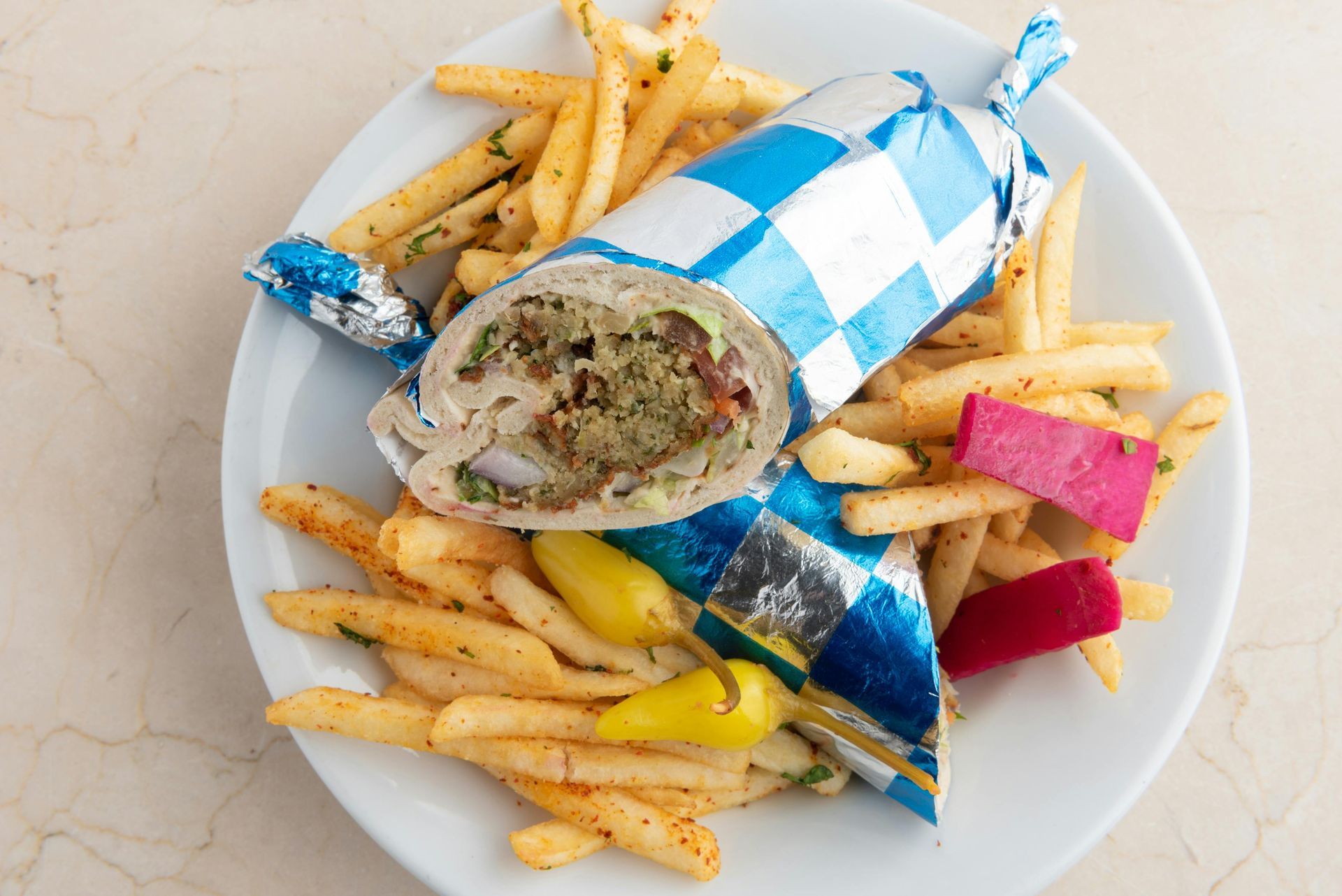 Falafel wrap and fries on a plate. The wrap is in checkered blue and white wrapping; fries are seasoned.