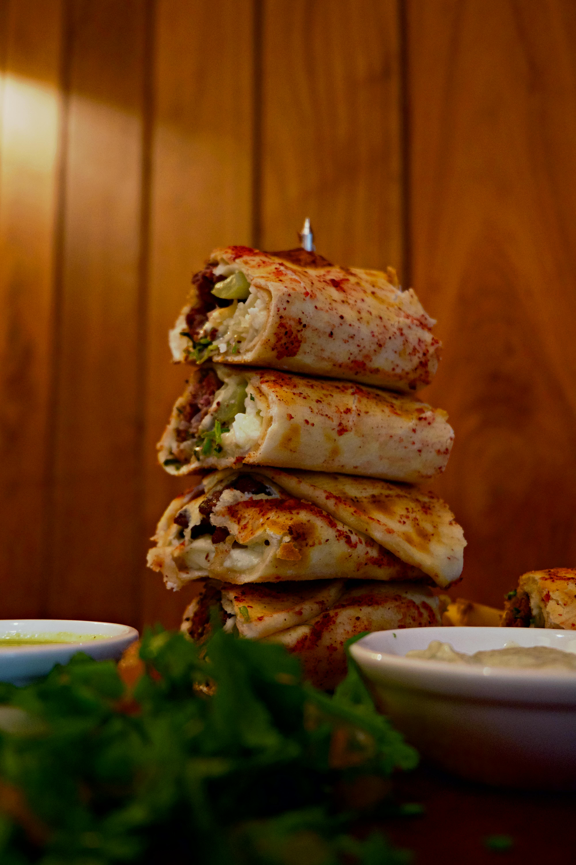 Stack of sliced wraps, skewered, with dipping sauces and parsley in front of a wood background.