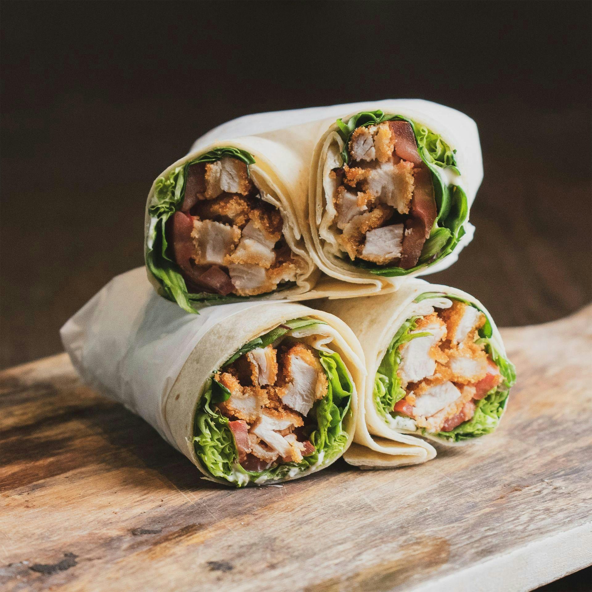 Three chicken wraps on a wooden board; contents include chicken, lettuce, and tomato.