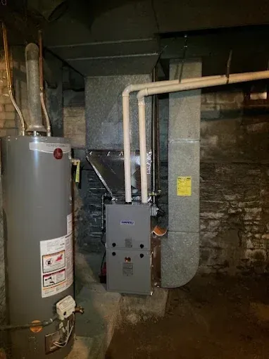 Water heater and furnace in a basement. Gray and copper pipes, metal ductwork, and a rough stone wall.