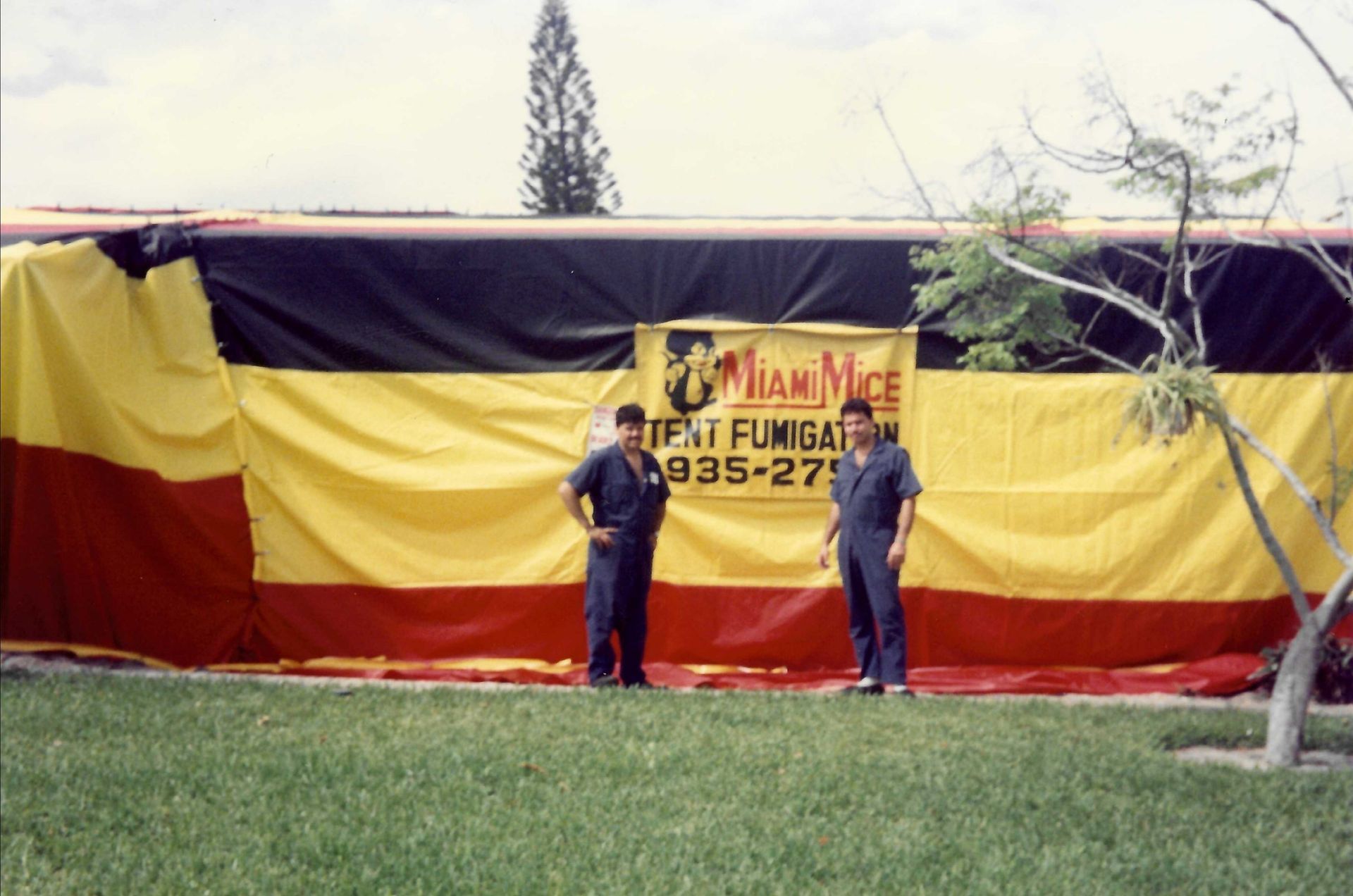 Two men in blue uniforms stand in front of a tented building, a sign reads 